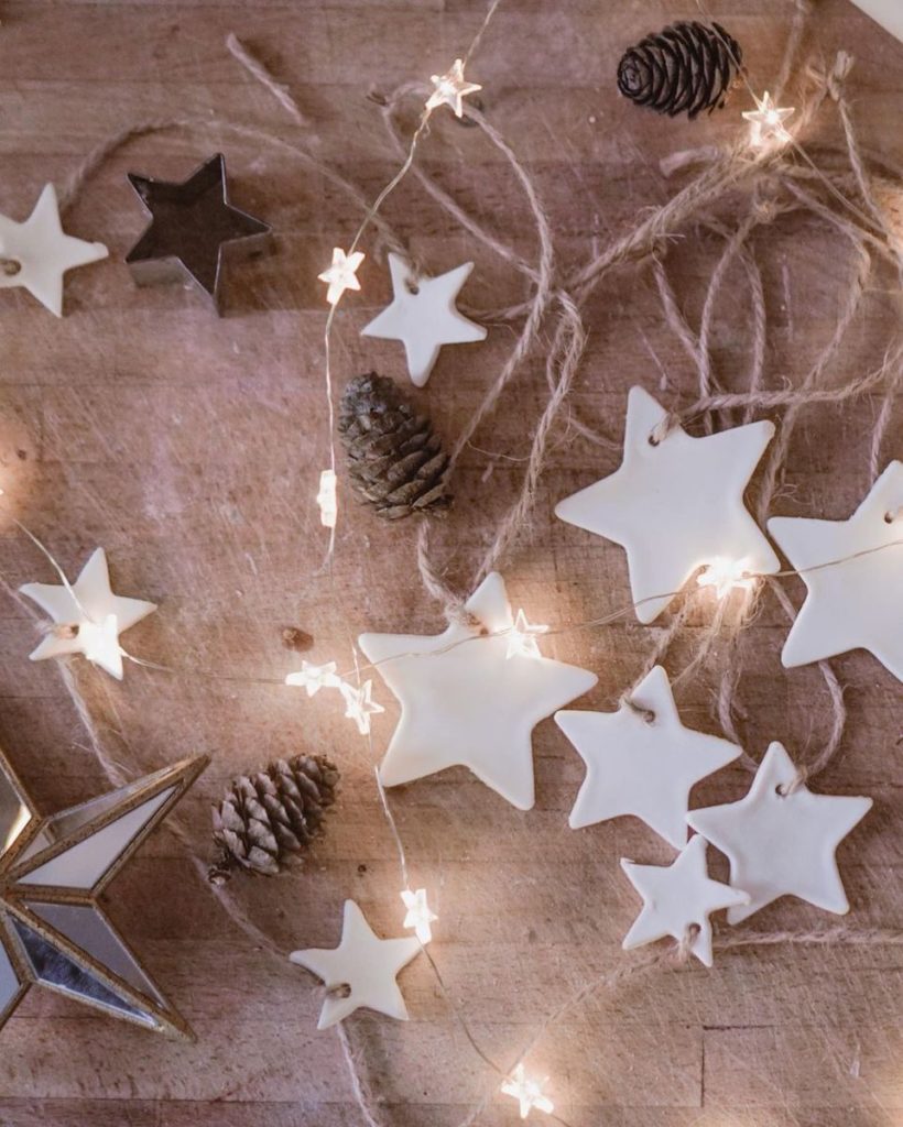 A flat lay image of white clay stars, Christmas lights and pinecones on a light wooden background.