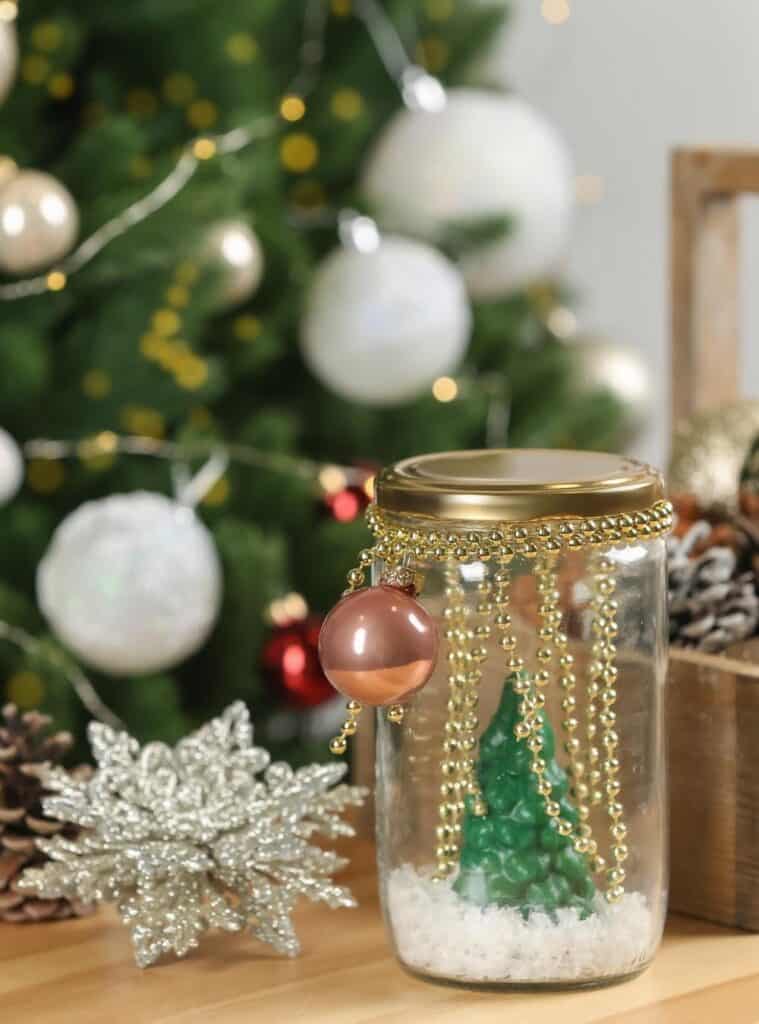 Upcycled DIY snow globe with mini trees and glitter inside a glass jar, sitting on a holiday table.