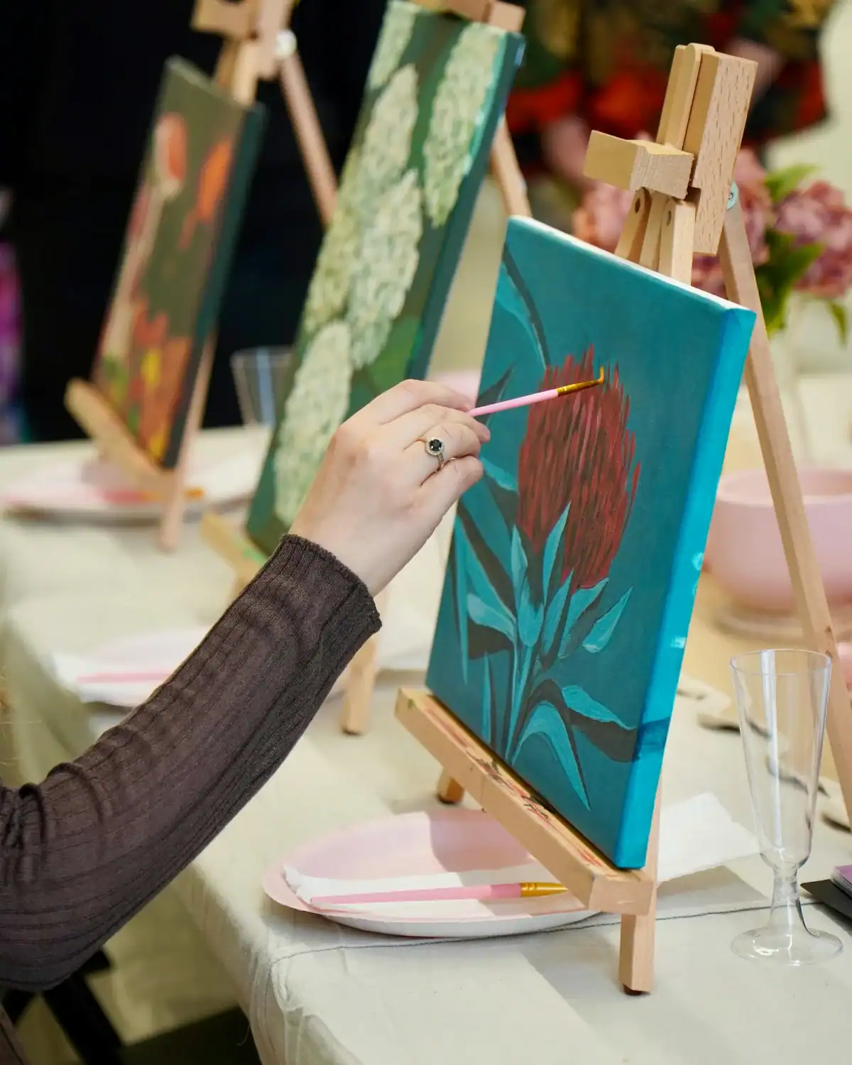 Person painting a red flower on a small canvas with acrylic paint and a wooden tabletop easel.