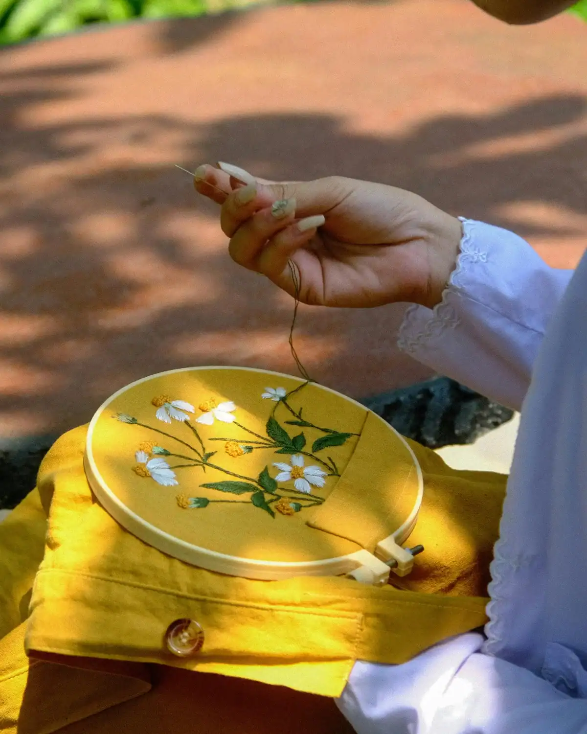 Person hand embroidering white and yellow flowers onto mustard fabric held in a wooden embroidery hoop.