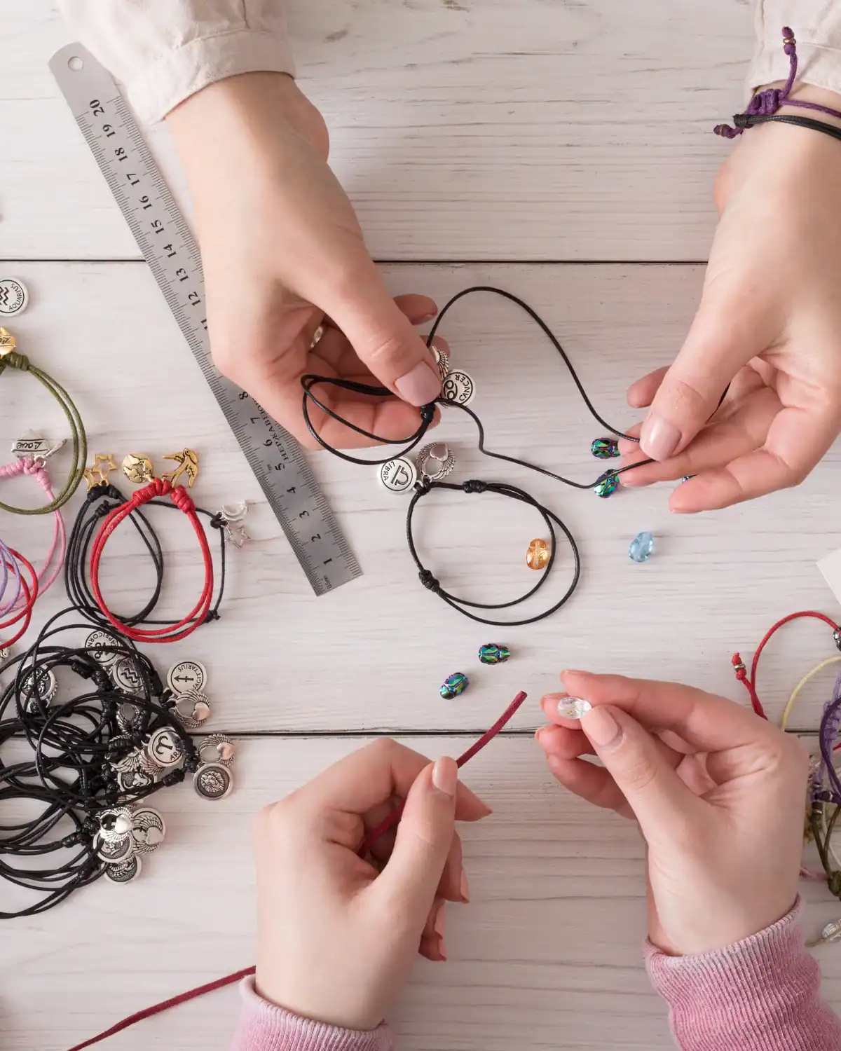 Hands making colorful cord bracelets with beads and metal charms on a craft table.