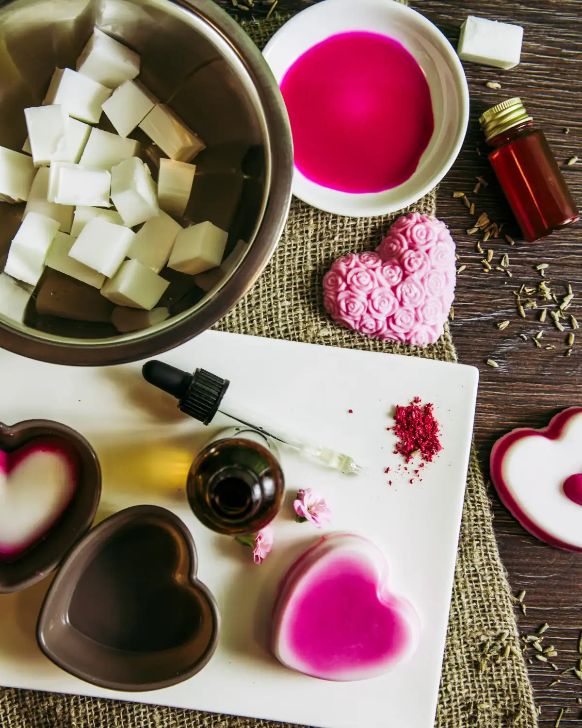 DIY melt-and-pour soap making setup with pink heart-shaped soap bars, soap base cubes, silicone molds, and botanical ingredients.