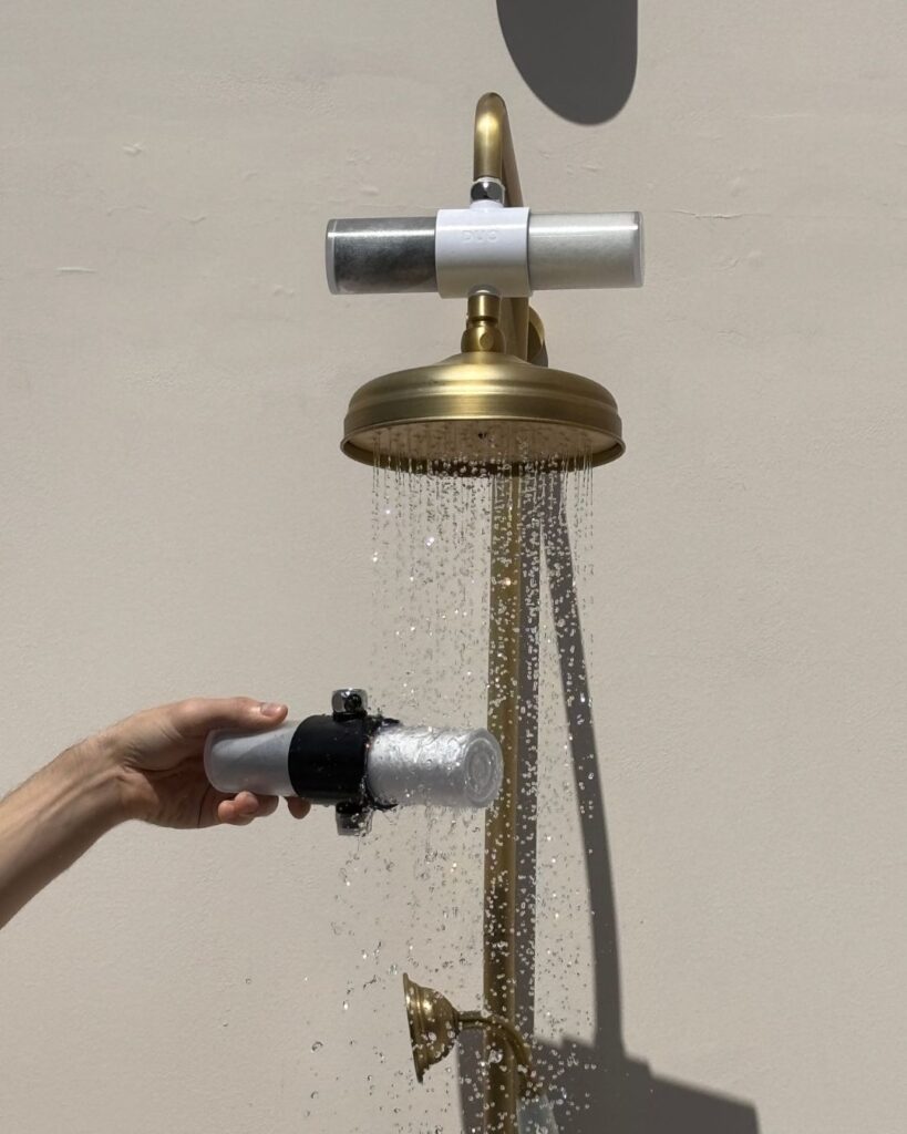 Weddell Duo shower filter installed above a brass showerhead, with a replaceable cartridge held under running water.