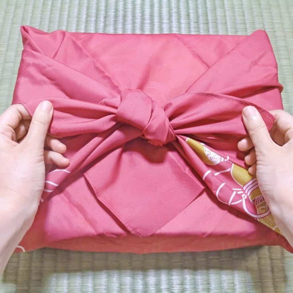 Hands tying a reusable Furoshiki fabric wrap around a Christmas gift as a non-toxic and sustainable gift wrapping alternative.