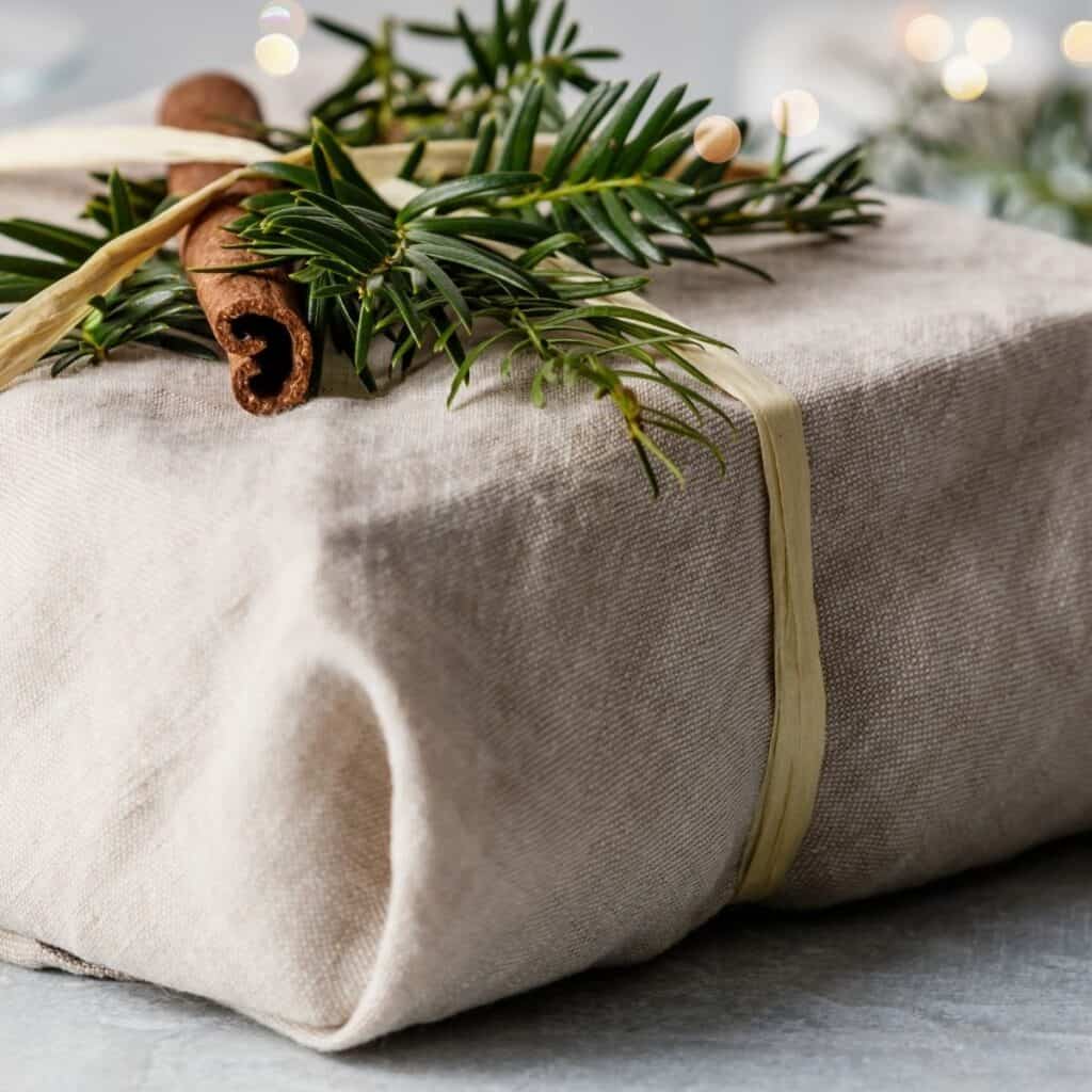 Christmas gift wrapped in natural linen fabric with evergreen sprigs and a cinnamon stick for reusable non-toxic Christmas gift wrapping.
