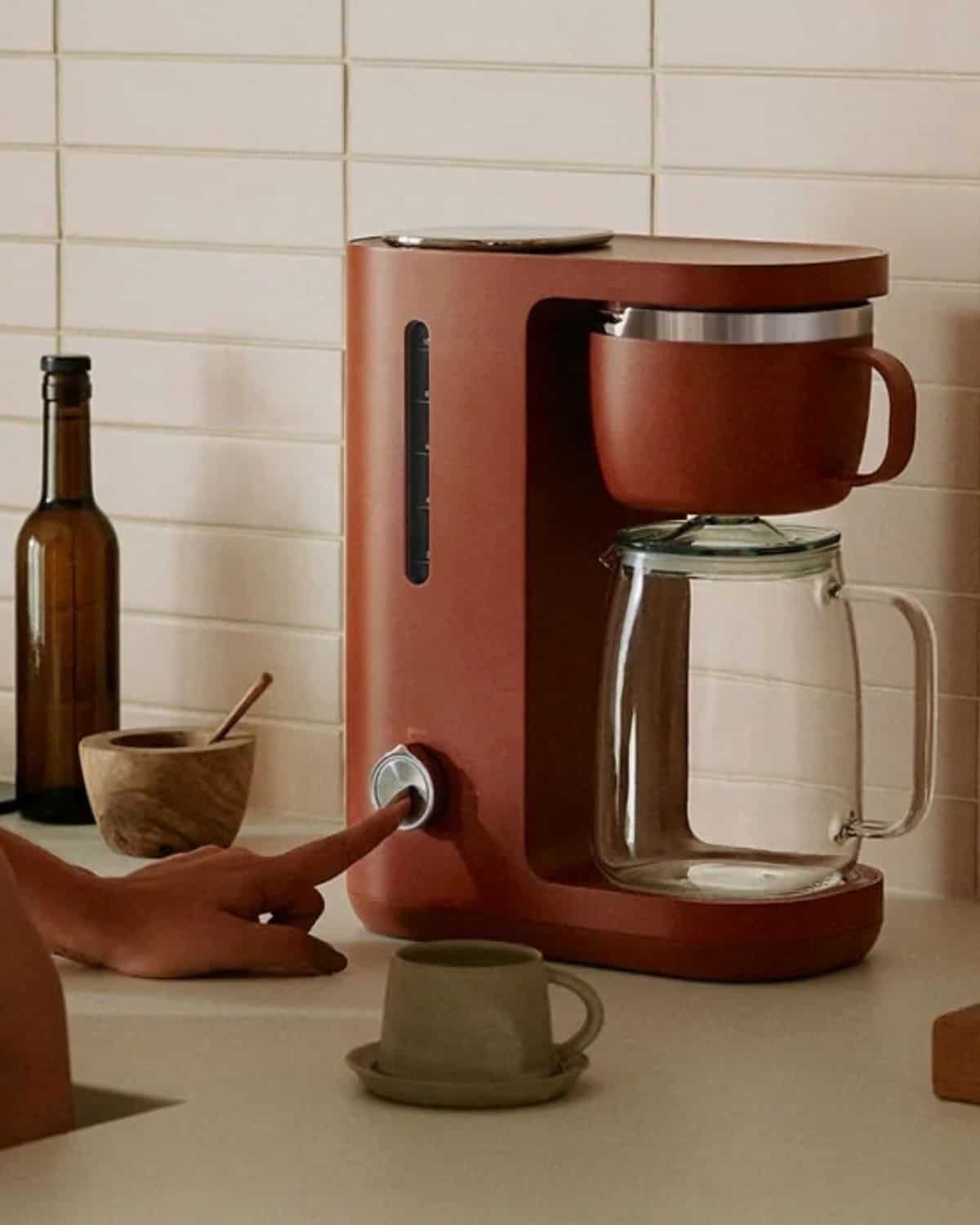 Hand adjusting the dial on a terracotta Caraway automatic coffee maker with a glass carafe