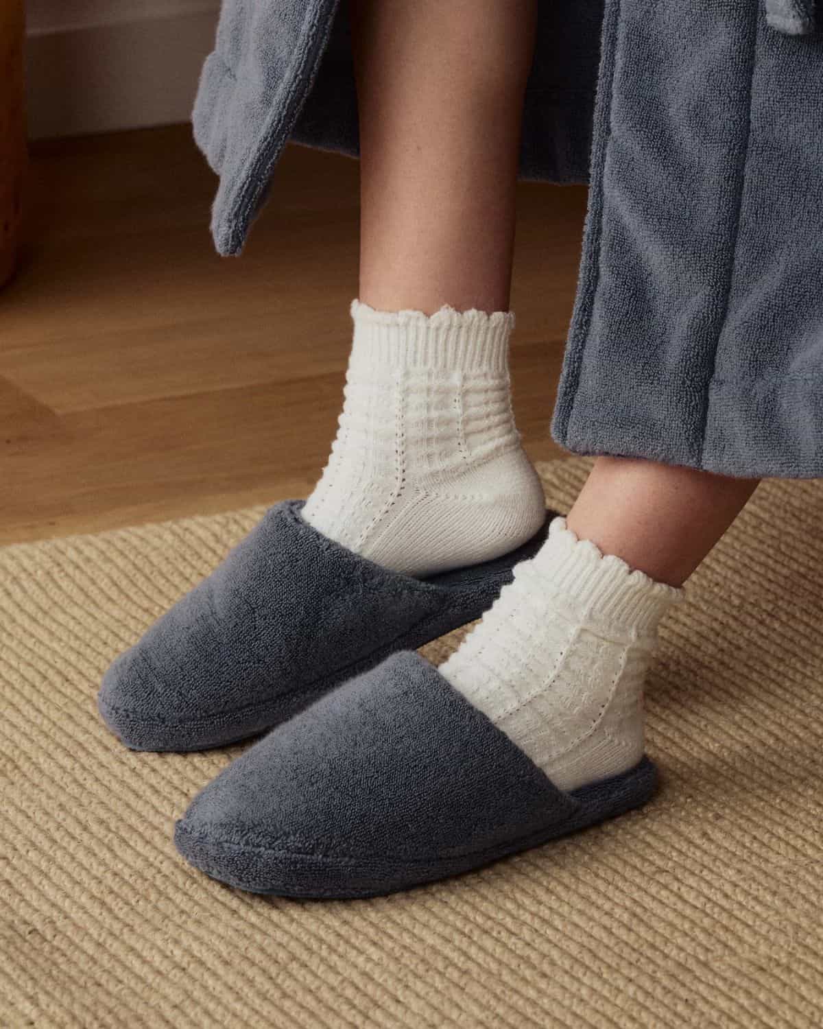 Dark blue Parachute Classic Plush Slippers worn with white knit socks and a blue robe on a beige woven rug.