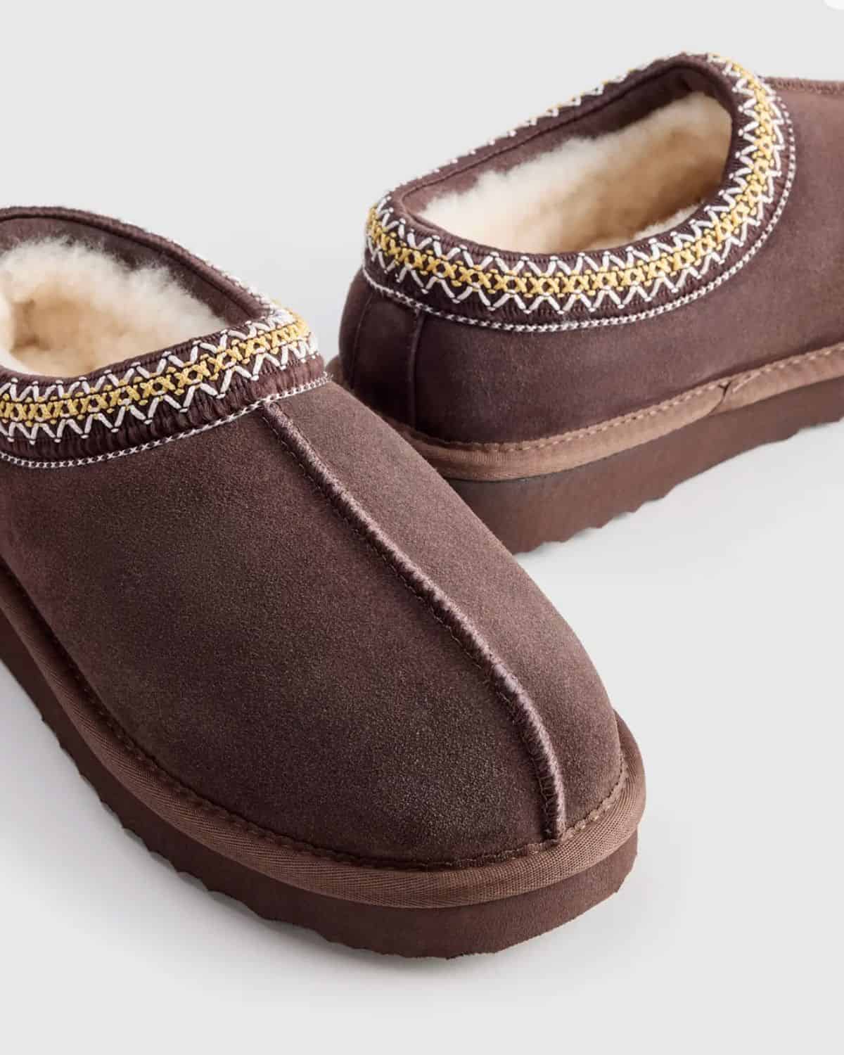 Brown Quince shearling clog slippers with cream lining, yellow and white embroidered trim, and thick brown soles.