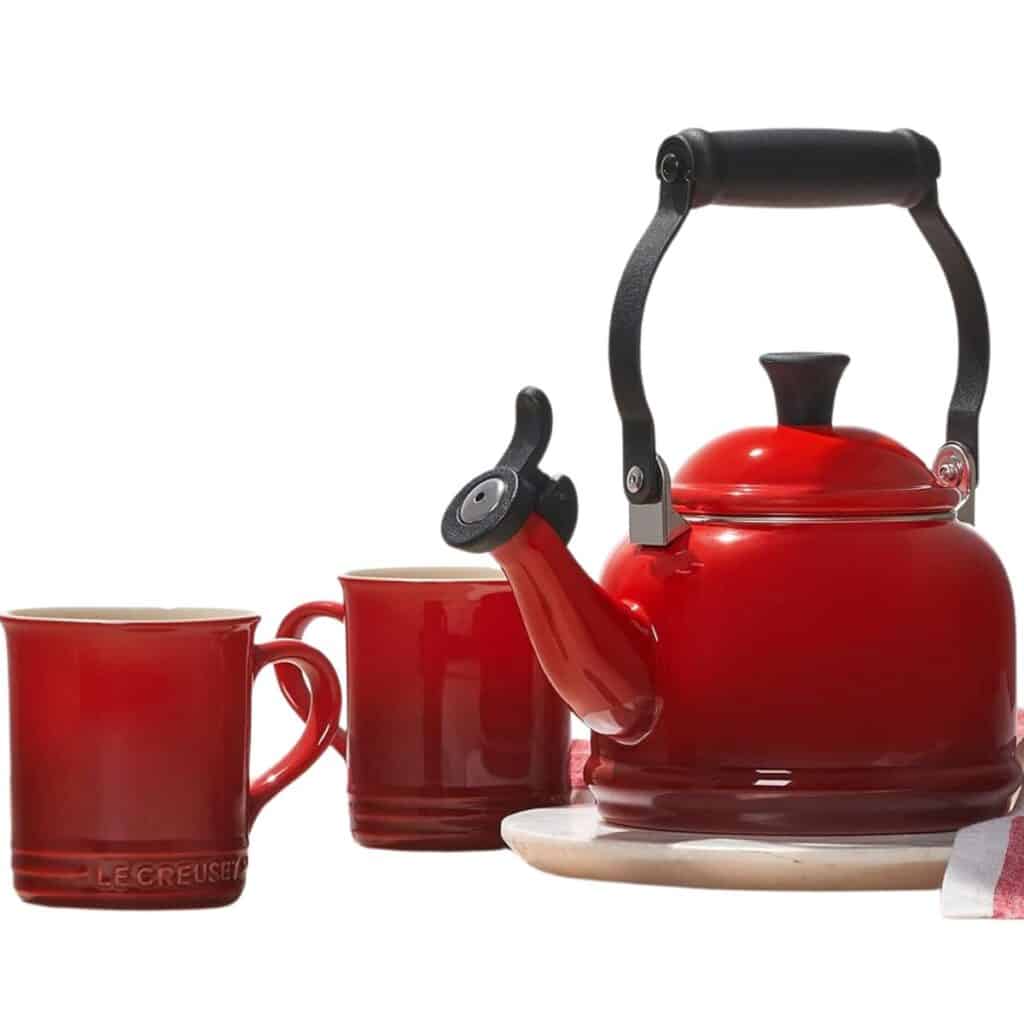 Red Le Creuset enamel-on-steel whistling tea kettle beside two matching red mugs
