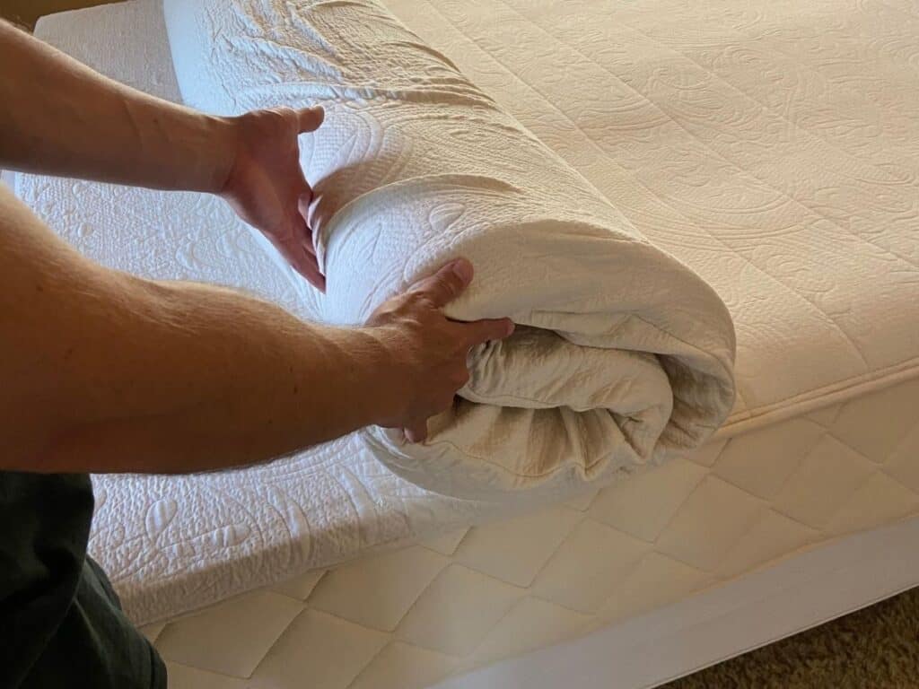 Hands rolling back the Happsy organic mattress topper to show its flexibility and the mattress underneath.